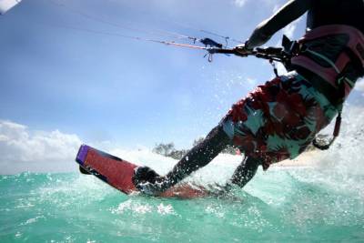A kitesurfer in floral shorts glides across turquoise water under a bright blue sky.