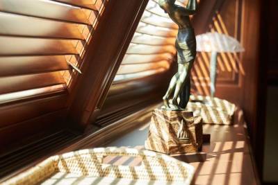 A woven tray sits in the foreground of a sunlit ledge, with a bronze female figurine standing before a window with closed wooden blinds.