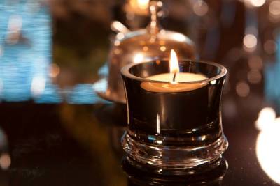 A lit tea light candle in a dark glass holder sits in the foreground against a blurred background of warm lights and a silver cloche.