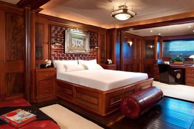 A luxurious bedroom with a large wooden bed and red bolster pillow in the foreground is set against a backdrop of rich wood-paneled walls and a built-in desk area.