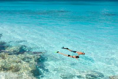 Two people snorkel in clear, shallow turquoise water over a rocky seabed.