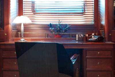 A sleek black office chair sits in the foreground before a polished wooden desk set against a background of rich mahogany walls and a window with blinds.