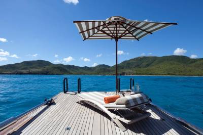 A striped beach umbrella and lounge chair sit on the wooden deck of a boat, overlooking a bright blue ocean and lush green mountains.