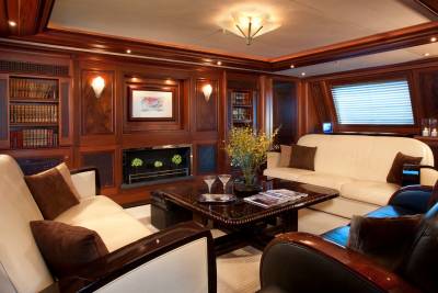 A luxurious wood-paneled lounge features white and brown leather sofas surrounding a dark coffee table in the foreground, with a fireplace and built-in bookshelves in the background.