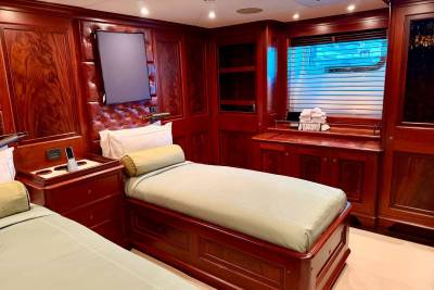 Two twin beds with light-colored linens sit in the foreground of a luxurious room featuring polished mahogany wood paneling and a window with blinds in the background.