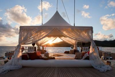 A luxurious white canopy bed with plush pillows sits on a wooden deck, set against a backdrop of calm ocean waters and distant mountains during a golden sunset.