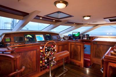 A polished wooden ship's bridge features a steering wheel and navigation screens in the foreground, with large windows revealing a cloudy sky in the background.
