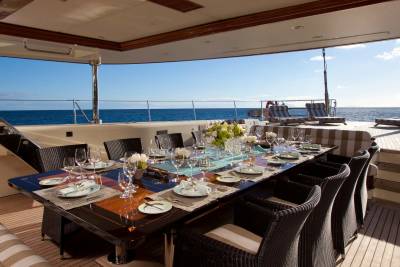 A lavishly set dining table with wicker chairs sits on the deck of a yacht, overlooking a calm blue ocean under a clear sky.