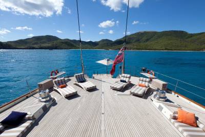A wooden yacht deck with lounge chairs and a red flag sits in the foreground, set against a backdrop of turquoise water and lush green mountains under a blue sky.