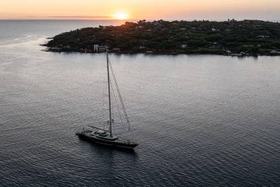 A sleek sailing yacht floats on calm dark waters in the foreground, with a lush, forested coastline and a setting sun in the background.