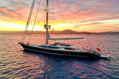 A sleek black sailing yacht floats on shimmering blue water against a backdrop of distant mountains and a vibrant sunset sky.
