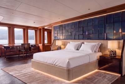 A luxurious bedroom features a large bed with white linens in the foreground, set against a backdrop of a textured blue wall and large windows overlooking a scenic waterfront.