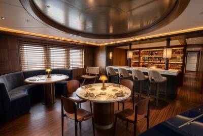 A luxurious dining area with round marble tables and blue seating sits in the foreground, while a sophisticated bar with high stools and backlit shelving fills the background.