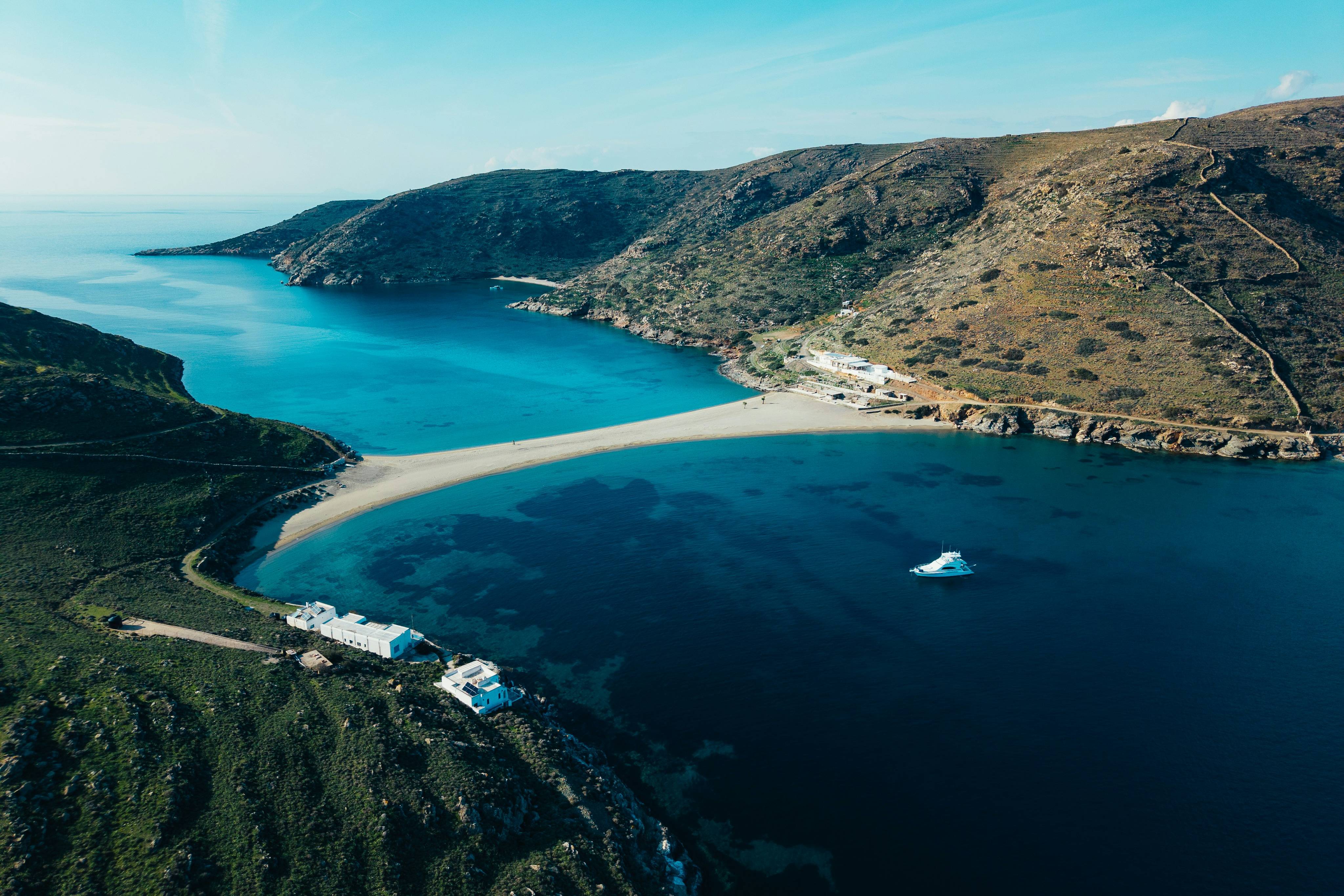 A narrow strip of sand connects two lush green hills, with a white boat sailing in the deep blue water.