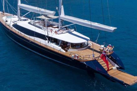 A luxurious dark blue yacht with white sails glides through crystal blue waters under a bright sky.