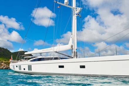 A sleek white yacht named Archelon sails through turquoise waters under a bright blue, cloud-dotted sky.