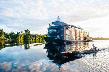 A man paddles a small canoe alongside the luxurious Aria Amazon cruise ship on a calm river.