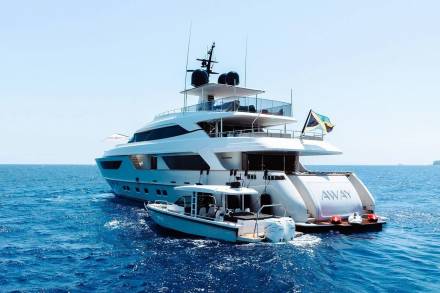 A sleek white superyacht named "AWAY" cruises through the vibrant blue ocean, trailed by a smaller tender boat.
