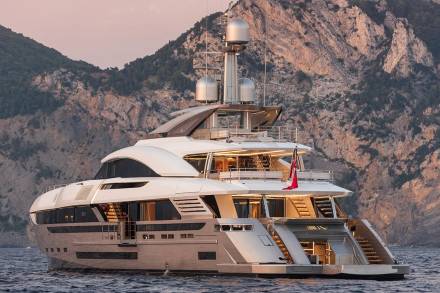 A luxurious white yacht glides through calm waters against a backdrop of rugged, sun-drenched mountains.