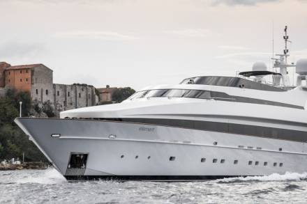A sleek white yacht named "element" glides through choppy waters past ancient buildings on a rocky coastline.