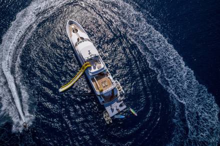 A luxurious yacht with a yellow slide creates swirling wake patterns on the deep blue ocean as a jet ski speeds nearby.