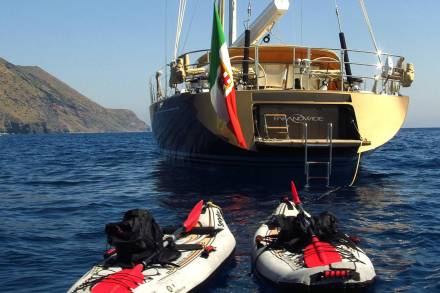Two inflatable kayaks with black backpacks bob in the blue ocean near a large golden yacht flying an Italian flag.