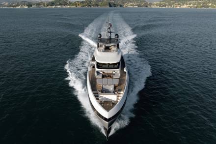 A sleek white yacht cuts through the dark blue water, leaving a foamy wake against a backdrop of green hills.