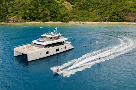 A luxurious catamaran anchors near a lush tropical island as people enjoy water sports in the crystal-clear blue ocean.
