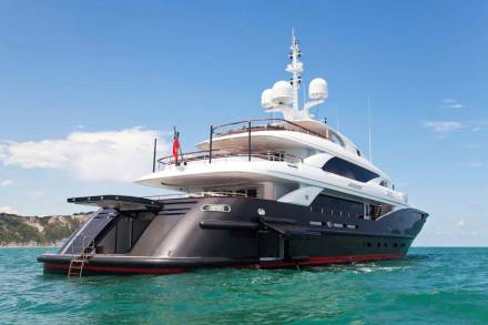 A luxurious dark hull yacht with multiple decks cruises on turquoise waters under a bright blue sky.
