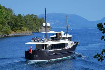 A sleek yacht cruises through a tranquil fjord surrounded by lush green hills under a clear blue sky.