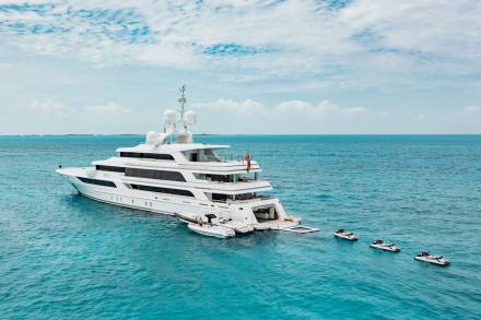 A luxurious white yacht cruises through crystal-clear turquoise waters, flanked by tender boats and jet skis.