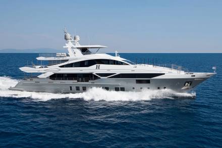A large, luxurious white yacht is speeding across the calm, blue waters of the ocean, with mountains visible in the distant background.