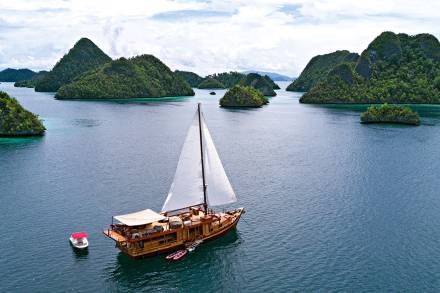 A majestic wooden sailboat glides through the turquoise waters of Raja Ampat, surrounded by lush, emerald islands.
