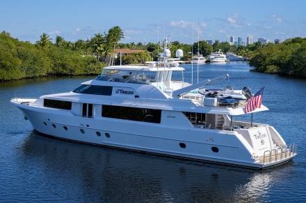 A large white yacht named "Tireless" cruises down a calm waterway lined with lush green trees.