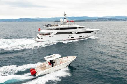 A sleek white yacht cruises the azure waters, trailed by a smaller speedboat under a cloudy sky.