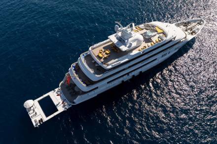 A luxurious superyacht with multiple decks and a swimming platform glides through the sun-drenched Mediterranean Sea.