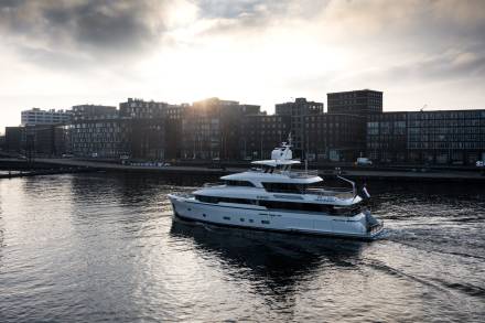 A sleek white yacht glides through the water with a modern city skyline in the background under a dramatic sky.