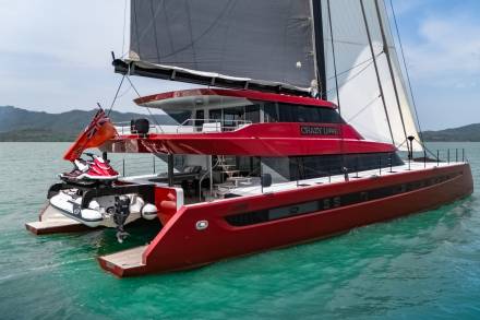 The striking red catamaran "Crazy Love" glides through turquoise waters, its sails catching the breeze under a bright sky.