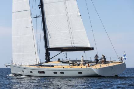 A sleek, modern sailboat glides through calm blue waters under a clear sky, its white sails full of wind.