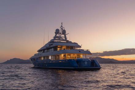 A luxurious yacht named Mosaïque glides across calm waters at sunset, its warm lights reflecting on the sea.