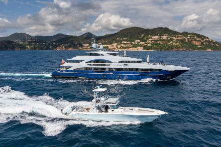 A sleek superyacht and a smaller chase boat cruise through the azure waters near a verdant, populated coastline.