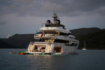 A luxurious yacht anchors in a serene bay as people enjoy the water and a smaller sailboat drifts nearby.