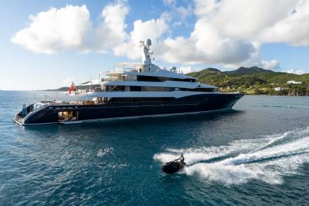 A person on a jet ski speeds through the turquoise water near a luxurious superyacht anchored offshore.