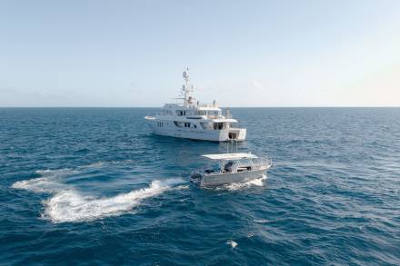 A sleek white yacht cruises the azure ocean, trailed by a smaller, agile boat creating a foamy wake.