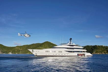 A sleek white helicopter approaches a luxurious superyacht anchored in a tropical bay.