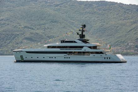 A majestic white superyacht adorned with colorful flags sails serenely on calm blue waters against a backdrop of lush green hills.