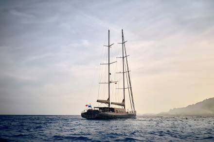 A majestic two-masted sailing yacht cuts through the deep blue ocean under a hazy sky.