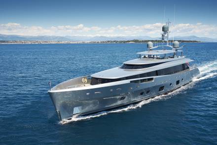 A sleek, modern yacht glides through the deep blue sea with a picturesque coastline and snow-capped mountains in the distance.