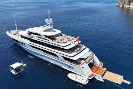 A luxurious superyacht with a jacuzzi and tender boat is anchored in the clear blue waters near a rocky coastline.