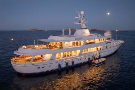 A luxurious yacht named Lady Beatrice is illuminated at dusk while anchored near a rocky coastline under a full moon.